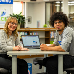 Staff member sits at table in the library with a student. A laptop is between them. Both are looking at the camera.