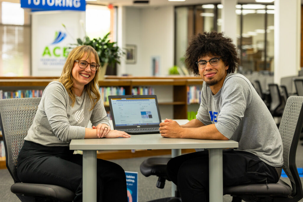 Staff member sits at table in the library with a student. A laptop is between them. Both are looking at the camera.