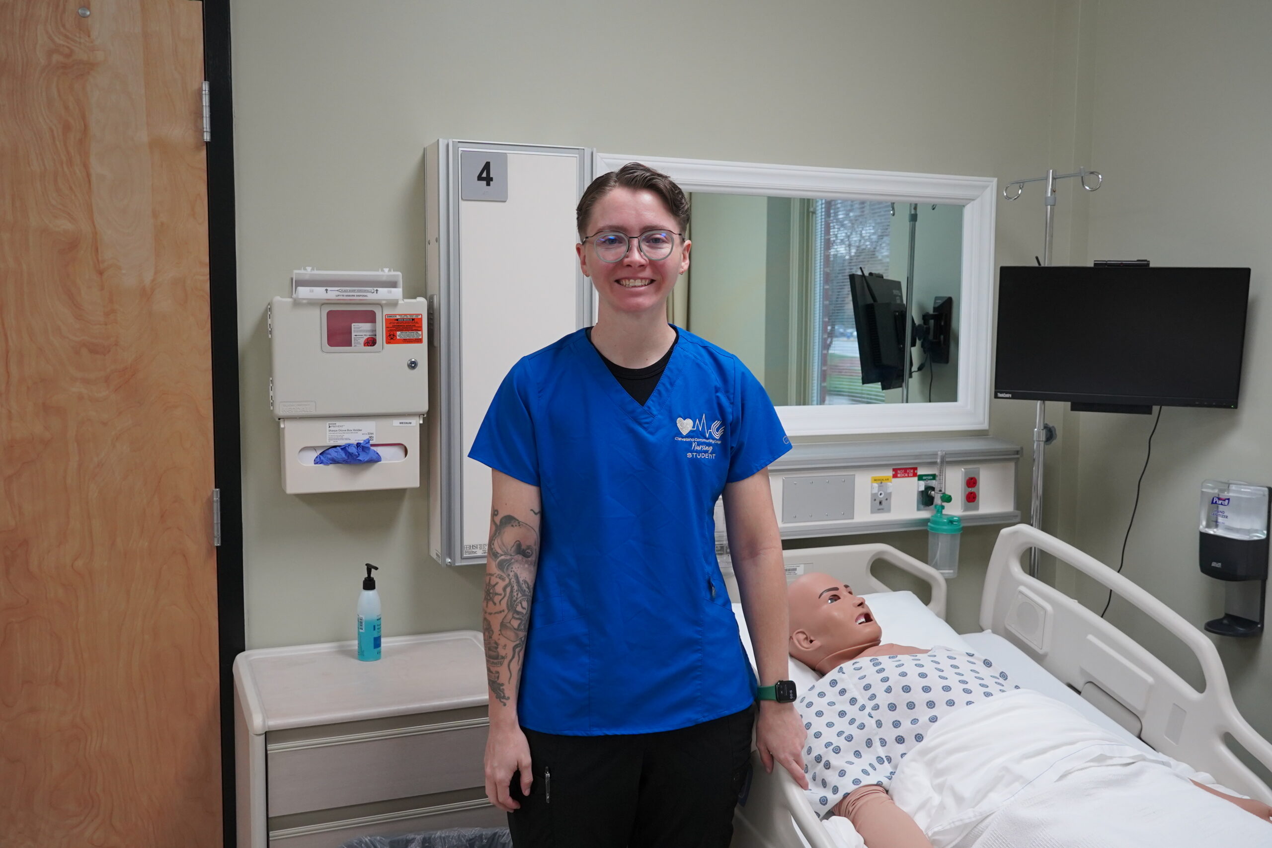 Mallory, wearing her nursing scrubs, stands in the simulation lab and smiles at the camera.