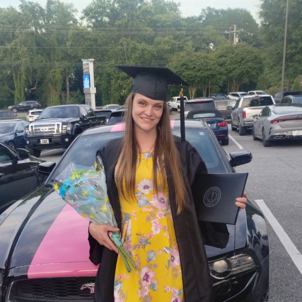 Miranda Parks in cap and gown showing off her diploma and holding flowers.